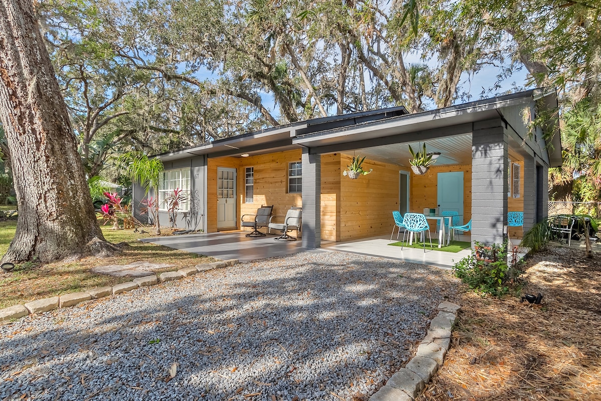 The exterior of the bungalow is set under mature oak trees, showcasing a charming combination of wooden and modern features. A spacious patio area is furnished with colorful outdoor chairs and a table, surrounded by landscaped gravel pathways and greenery.