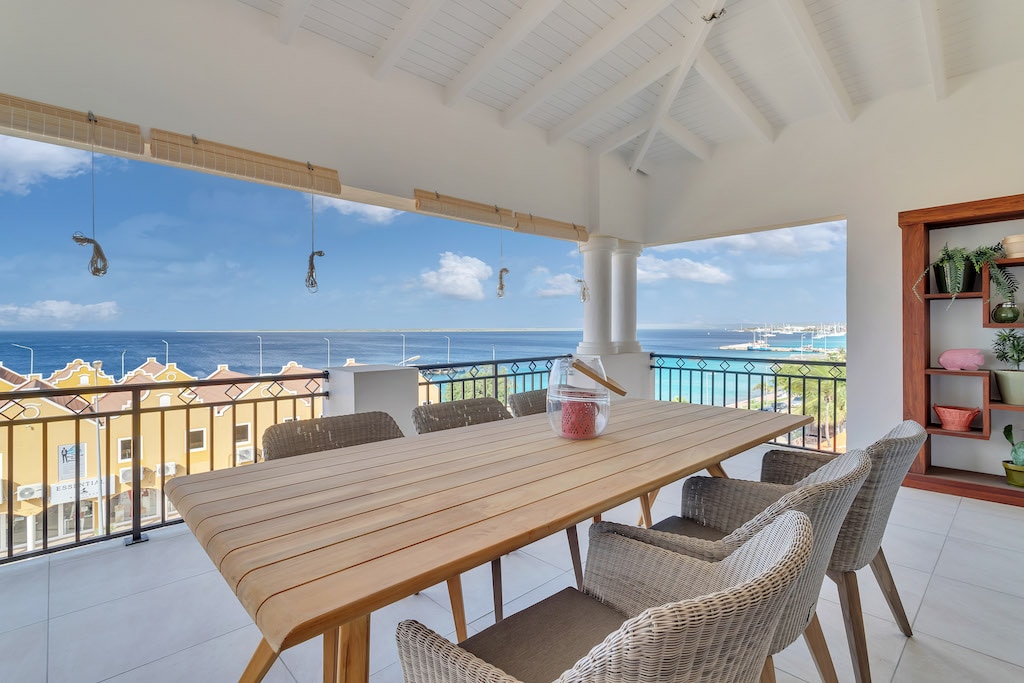 A spacious balcony is showcased with a large wooden dining table surrounded by six woven chairs. The area offers panoramic views of the ocean and shoreline, with the sky appearing bright and clear. Decorative elements are subtly integrated into the space.