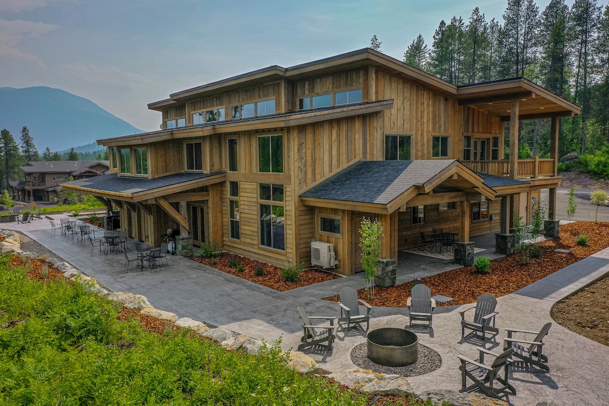The exterior of the condo features natural wood siding, large windows, and a welcoming patio area. A fire pit is surrounded by seating, offering a spot for evening gatherings. Lush landscaping and surrounding trees complement the serene mountain backdrop.