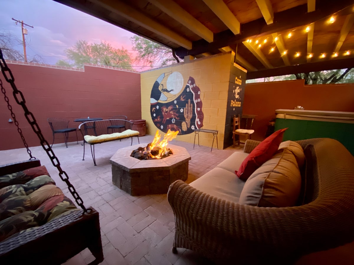 An inviting outdoor space features a circular stone fire pit, surrounded by comfortable seating and a snug sofa with throw pillows. Warm lights hang overhead, illuminating a mural on the wall, enhancing the tranquil ambiance of the patio at dusk.