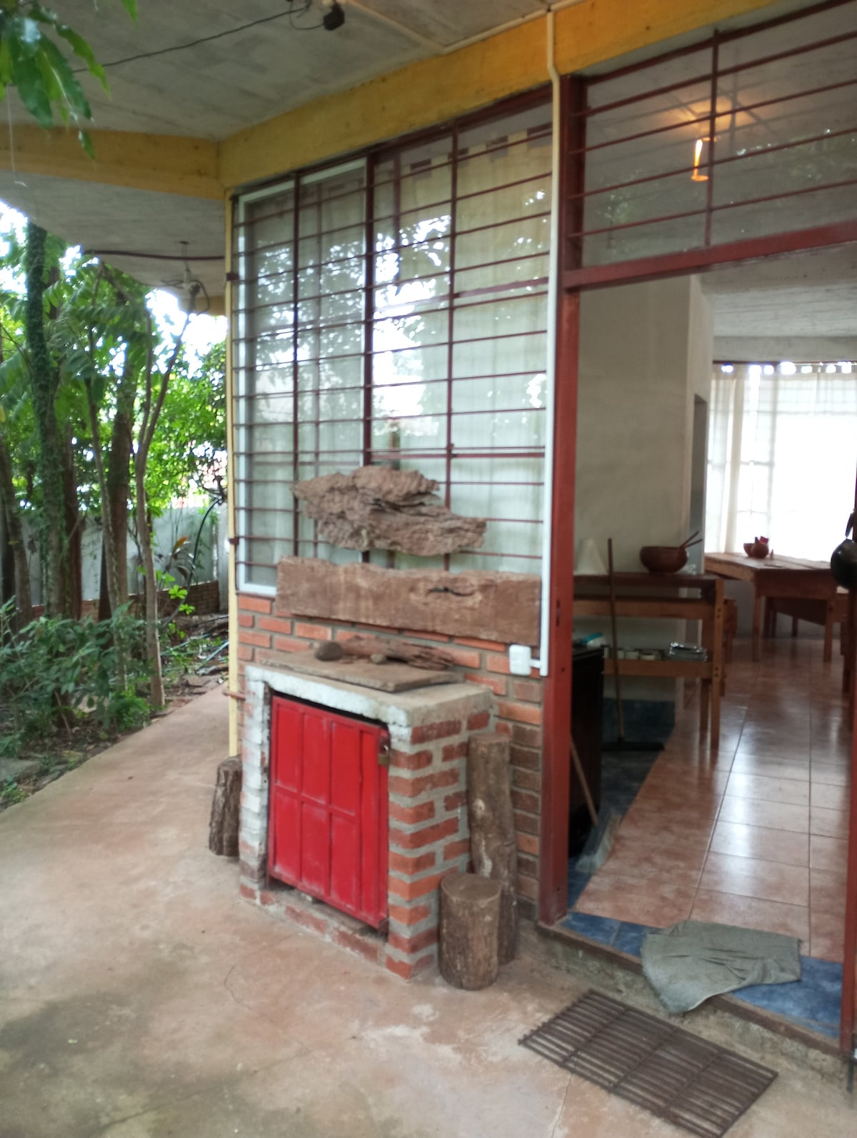 An outdoor area features a rustic brick barbecue with a red door. Surrounding greenery offers a natural setting, while a large window with metal bars allows light to enter the adjacent indoor space, where wooden furniture can be seen.