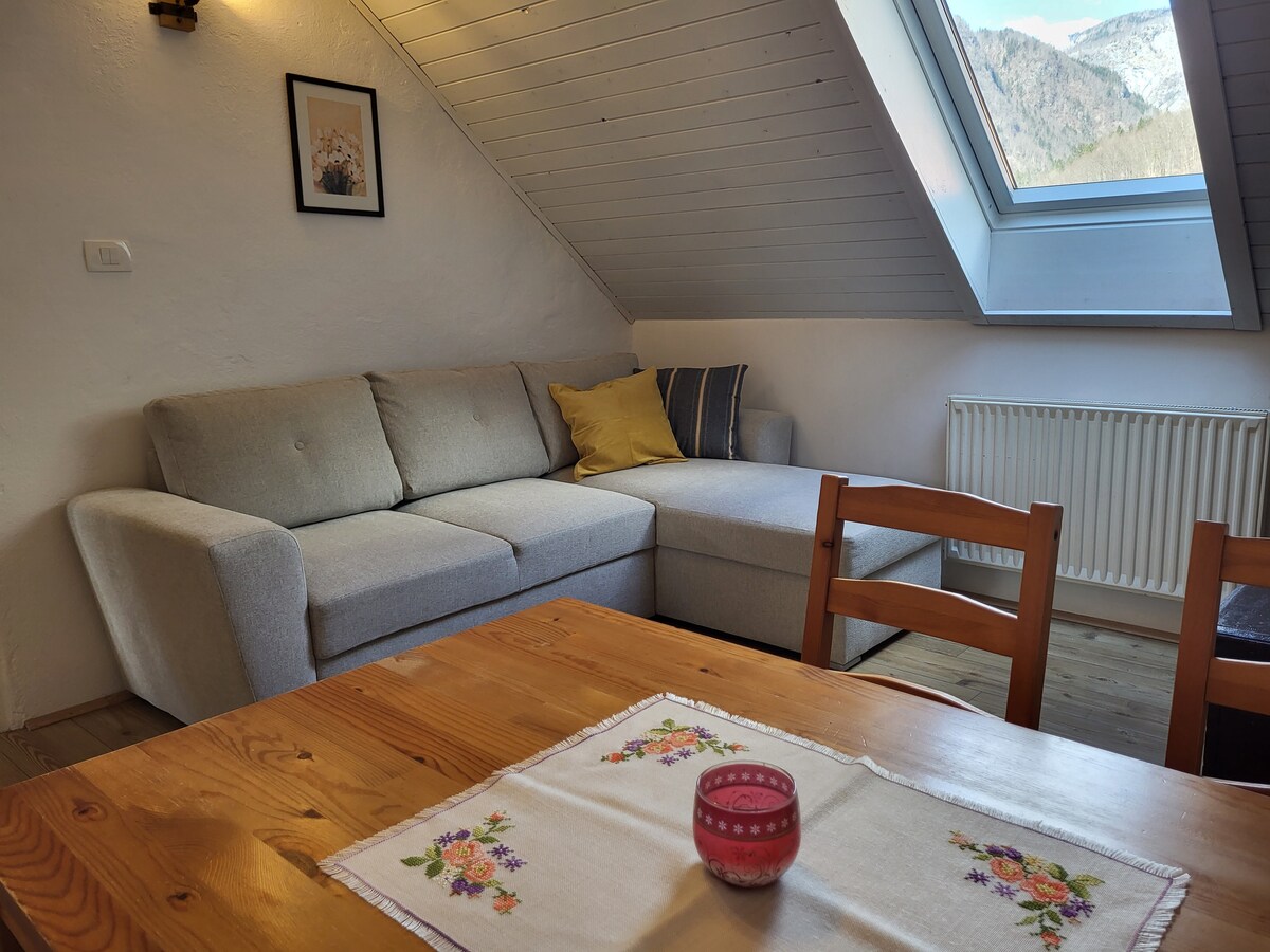 A comfortable seating area is shown with a light gray sectional sofa adorned with a yellow throw pillow. A wooden dining table featuring a floral tablecloth and a pink candle sits nearby. A large skylight offers views of the mountains in the background.