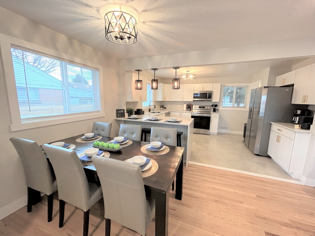 A dining area is positioned next to an open kitchen, featuring a large wooden table set for eight with modern grey chairs. Soft lighting from hanging fixtures illuminates the space, while large windows allow natural light to fill the room. Stainless steel appliances are visible in the kitchen.