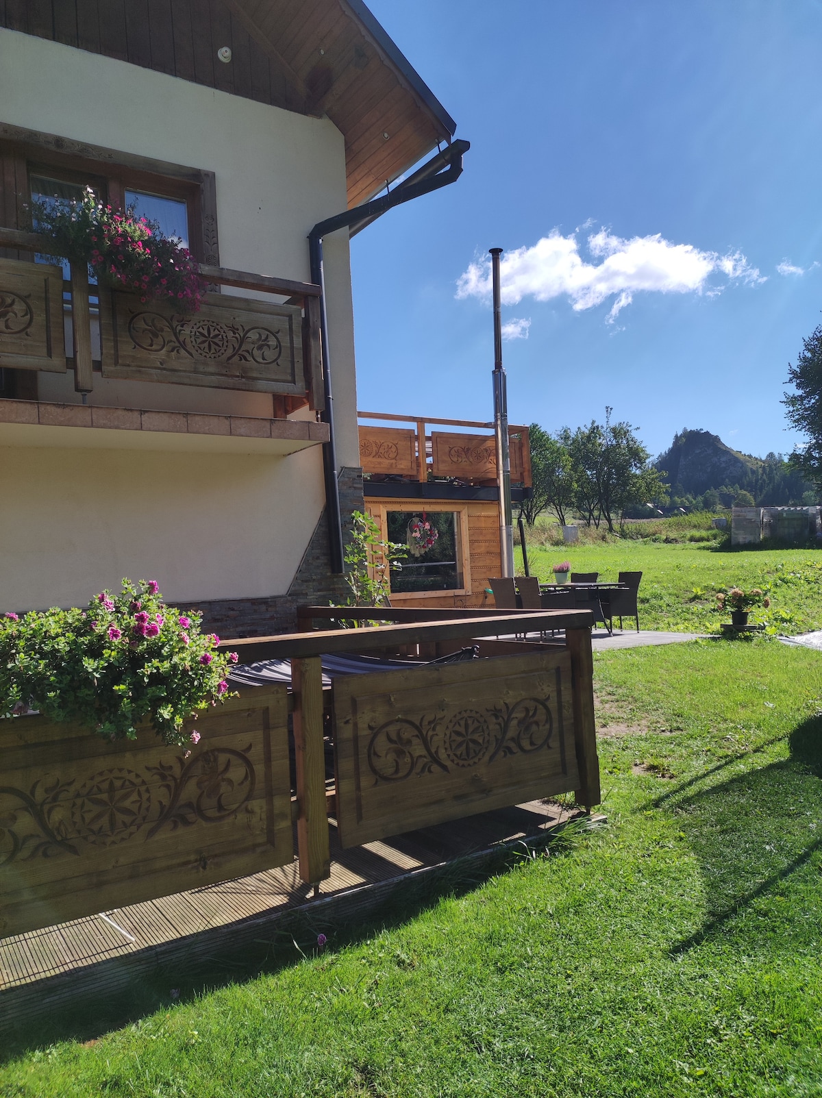 The exterior of a wooden cottage is presented, showcasing flower-filled balcony planters. Sunlight illuminates the surrounding greenery, enhancing the inviting outdoor space, which features seating arrangements. A nearby structure is visible, creating an atmosphere of rustic comfort amidst the natural landscape.