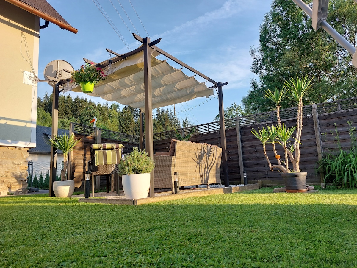 A shaded outdoor seating area is featured, with a pergola covered in fabric and adorned with hanging plants. A relaxed seating arrangement invites comfort, while neatly trimmed grass surrounds decorative potted plants and a basketball hoop stands nearby.