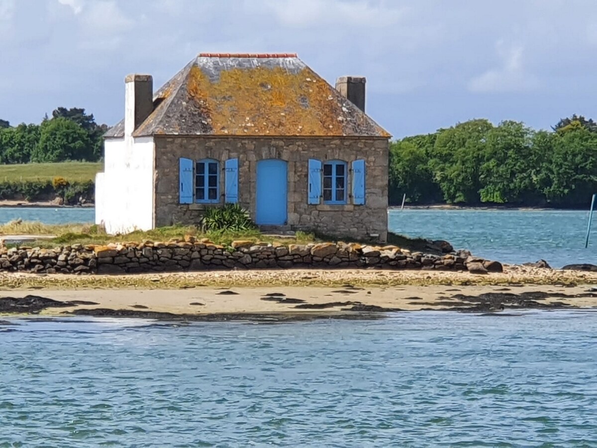 A charming stone house sits on a small island, surrounded by water. The structure features light blue shutters and a central blue door. A sandy shoreline leads to the house, and green trees can be seen in the background, blending with the coastal scenery.