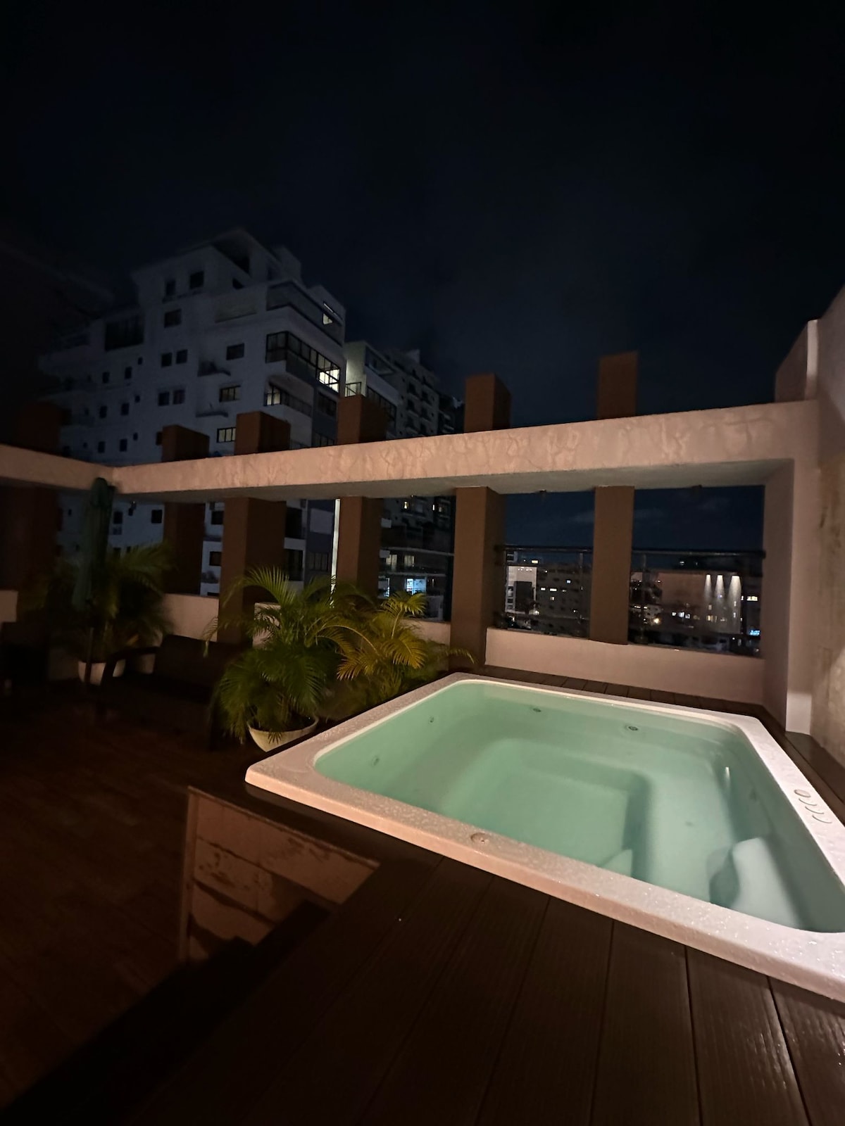 A rooftop terrace features a hot tub surrounded by wooden decking and potted plants. The city skyline is visible in the background, with ambient lighting casting a soft glow over the space at night.