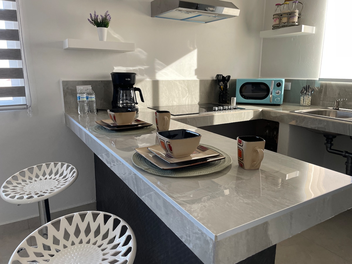 A modern kitchenette is showcased, featuring a sleek marble countertop. Two decorative bowls and a coffee maker are positioned on the counter, with a small vintage microwave nearby. A pair of white stools offers seating, while light streams in through the window with striped shades.