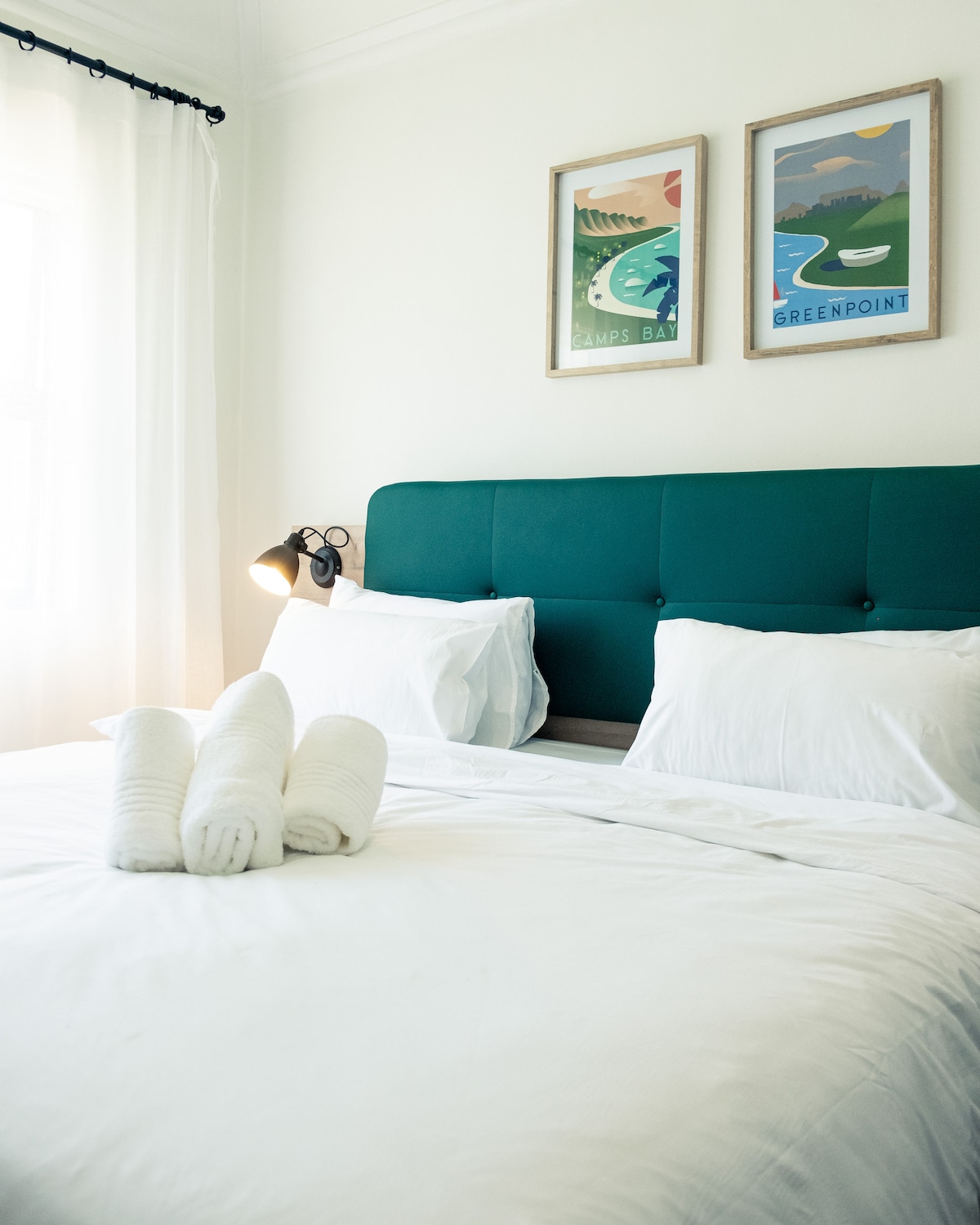 A spacious bed is adorned with crisp white linens and accompanied by neatly rolled towels. Above, two framed art pieces feature serene landscapes. Natural light enters from a window adorned with sheer white curtains, creating a bright and relaxing ambiance.