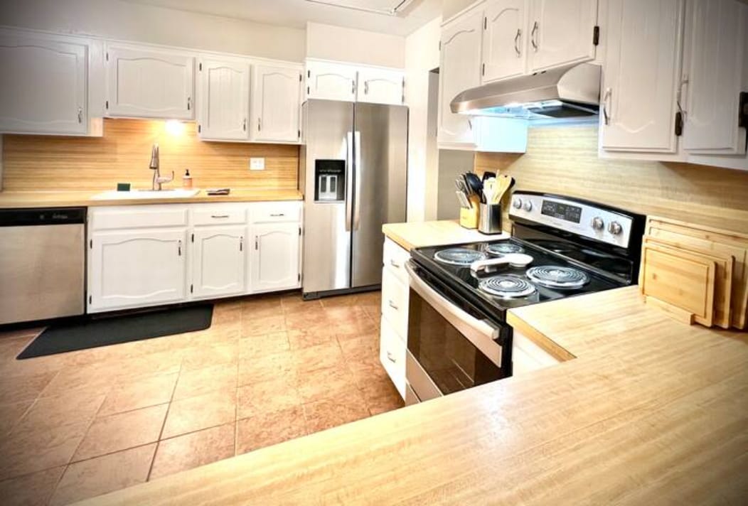 A spacious kitchen features modern stainless steel appliances, including a refrigerator and oven. White cabinetry offers ample storage, and a light-colored countertop provides a functional workspace. Cookware and utensils are neatly arranged, ensuring convenience for meal preparation.