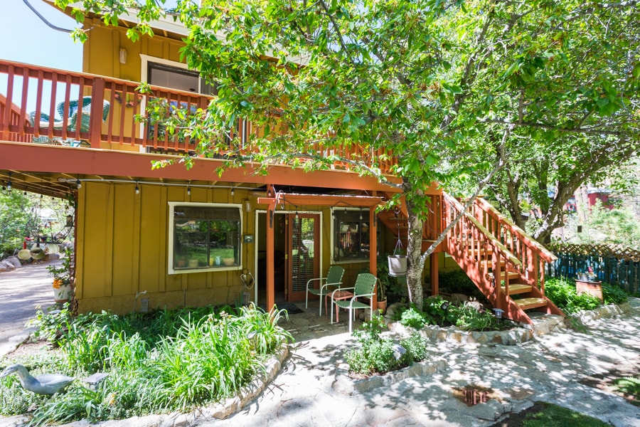 An inviting entrance to the Hidden Treasure Suite is surrounded by lush greenery and colorful landscaping. A staircase leads to the upper level, with a welcoming patio area featuring two chairs beside a well-maintained path. Natural light filters through large windows, enhancing the connection with the outdoors.
