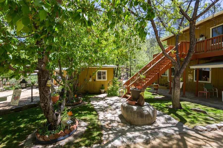 A lush outdoor area is surrounded by greenery and trees, featuring a well-maintained path leading to the entrance of the suite. A set of stairs is visible, providing access to the upper level. Decorative planter boxes and flowers add color to the landscape.