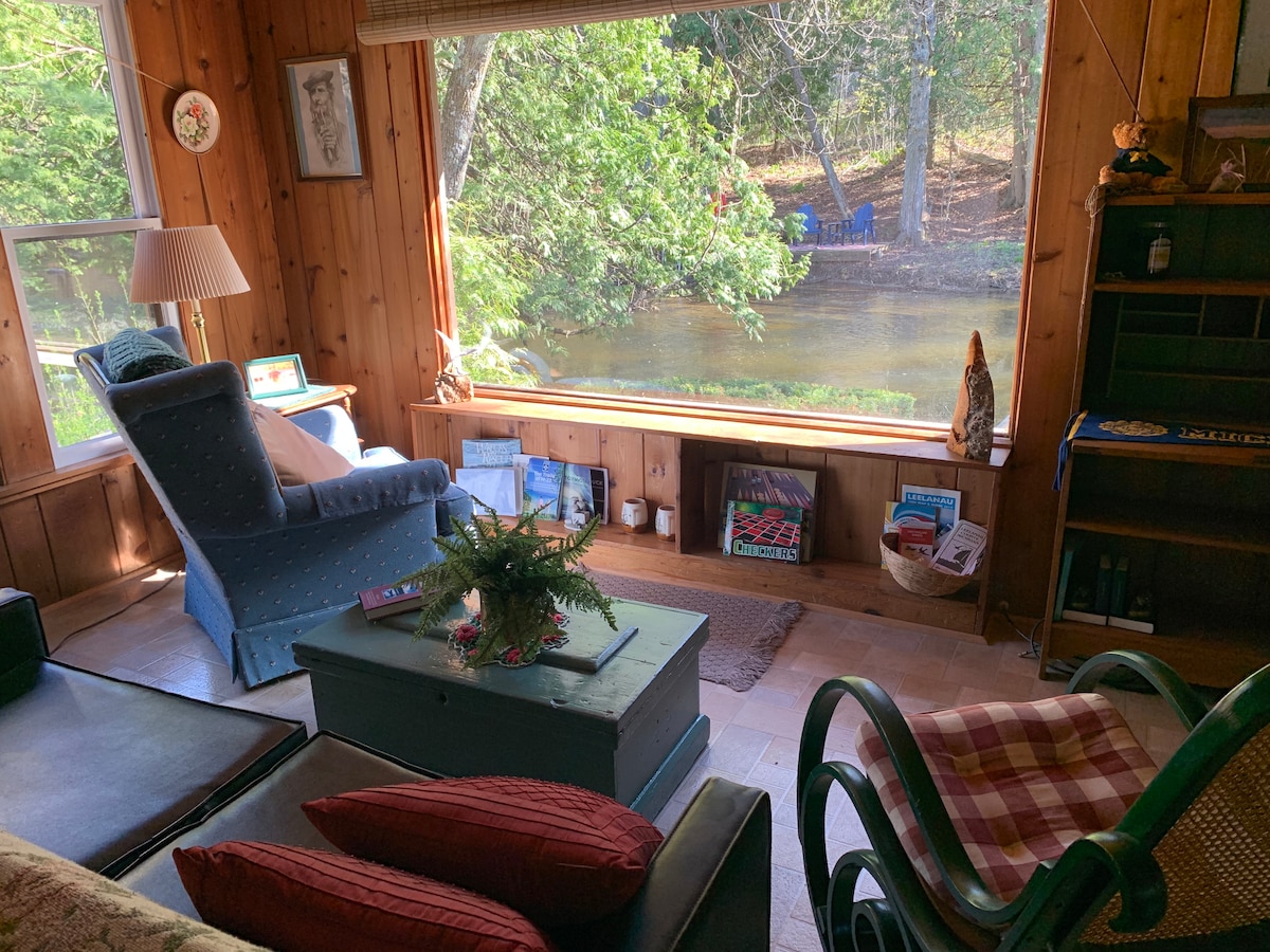 A cozy seating area is highlighted by a large window overlooking the Boardman River. Natural light filters in, illuminating wooden paneling and comfortable furnishings, including a blue armchair and a green rocking chair. A small coffee table rests in the center, adorned with decorative elements.