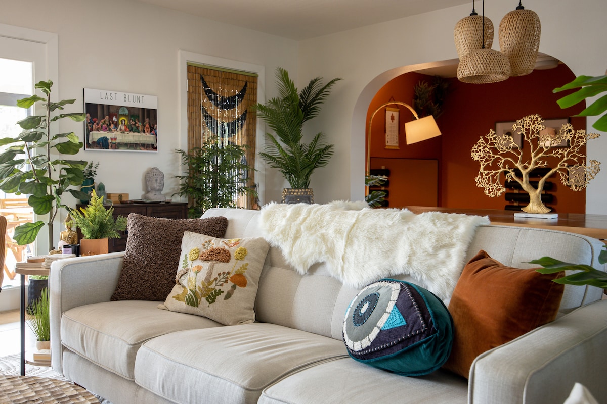A light-filled living room is showcased with a comfortable sofa adorned with decorative pillows. A large plant and a woven decorative piece enhance the cozy ambiance. Accent lighting and natural elements create a serene and inviting atmosphere, blending modern design with boho aesthetics.