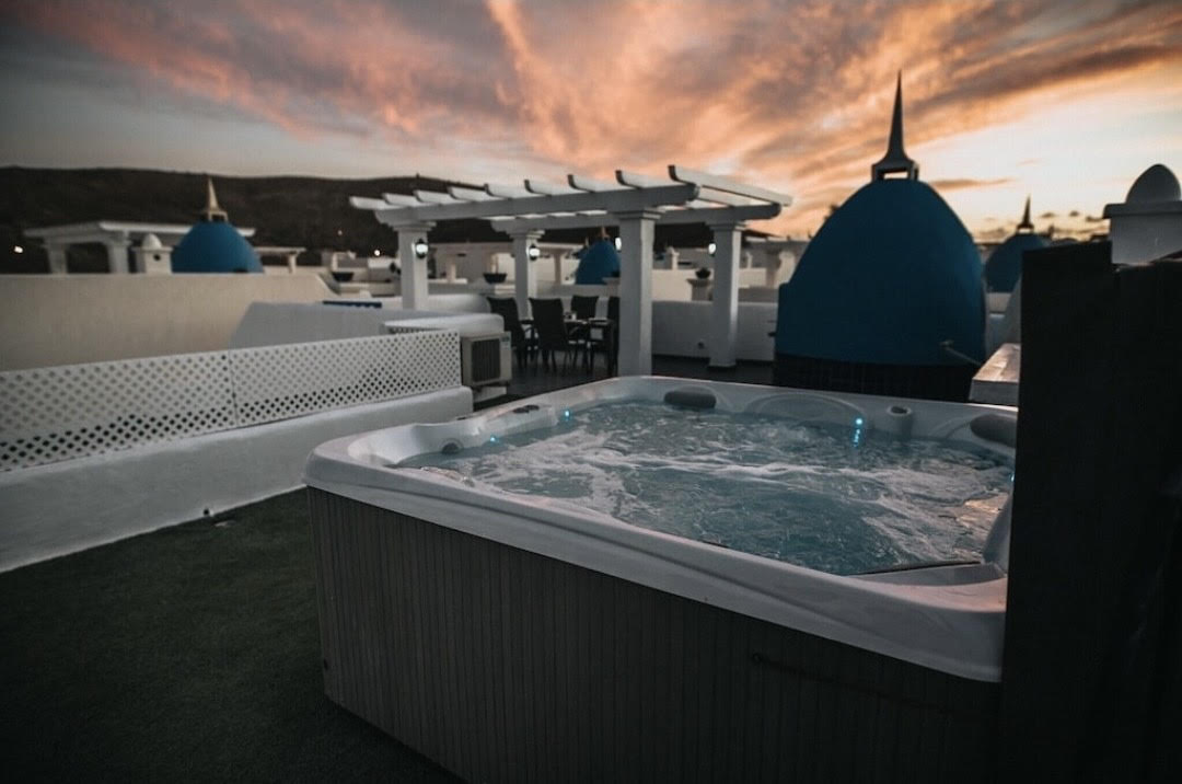 A heated hot tub is positioned on the roof terrace, surrounded by a green lawn. The area features a pergola, and the vibrant sky displays hues of orange and pink, adding a serene backdrop to the luxurious setting.