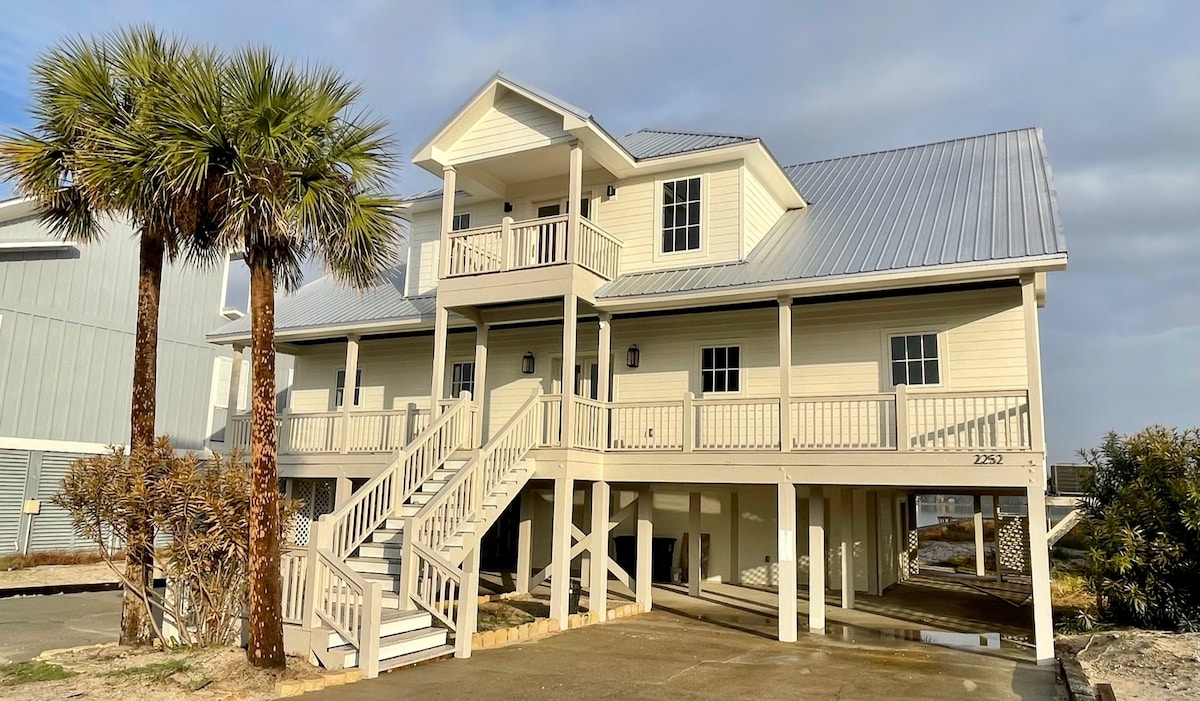A two-story house showcases a light-colored exterior with a metal roof. Multiple balconies and a front porch are visible. The entrance features stairs leading to the front door, while palm trees and landscaped areas enhance the setting. Space is provided under the house for parking.