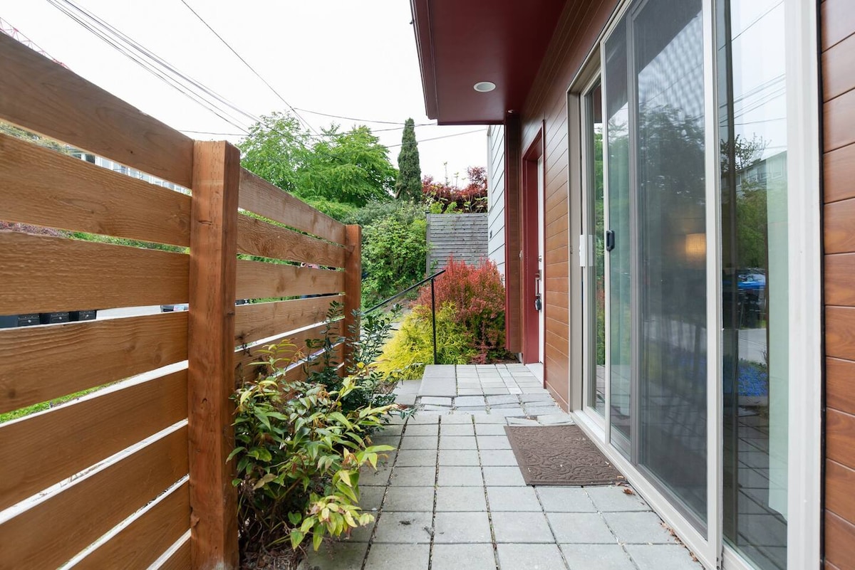 A private entrance is framed by a wooden fence and lush greenery. The pathway is paved with square tiles, leading to sliding glass doors that open to the guest suite. Steps are visible, providing access to the entryway.