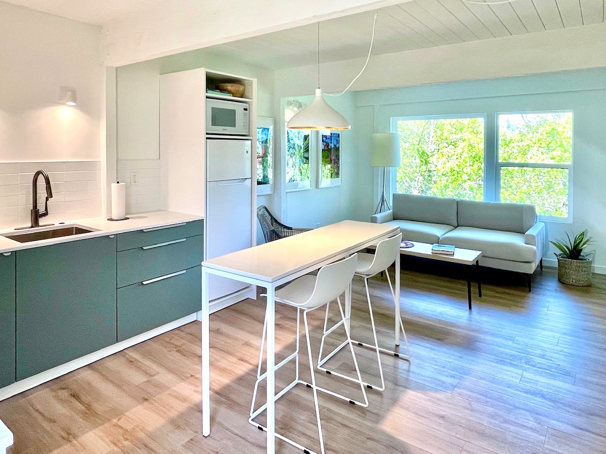 An open-concept kitchen and living area features a light, airy atmosphere with large windows allowing natural light to illuminate the space. A compact dining table for two sits next to the kitchen, which is equipped with modern appliances and sleek cabinetry. A cozy sofa adds to the inviting environment.