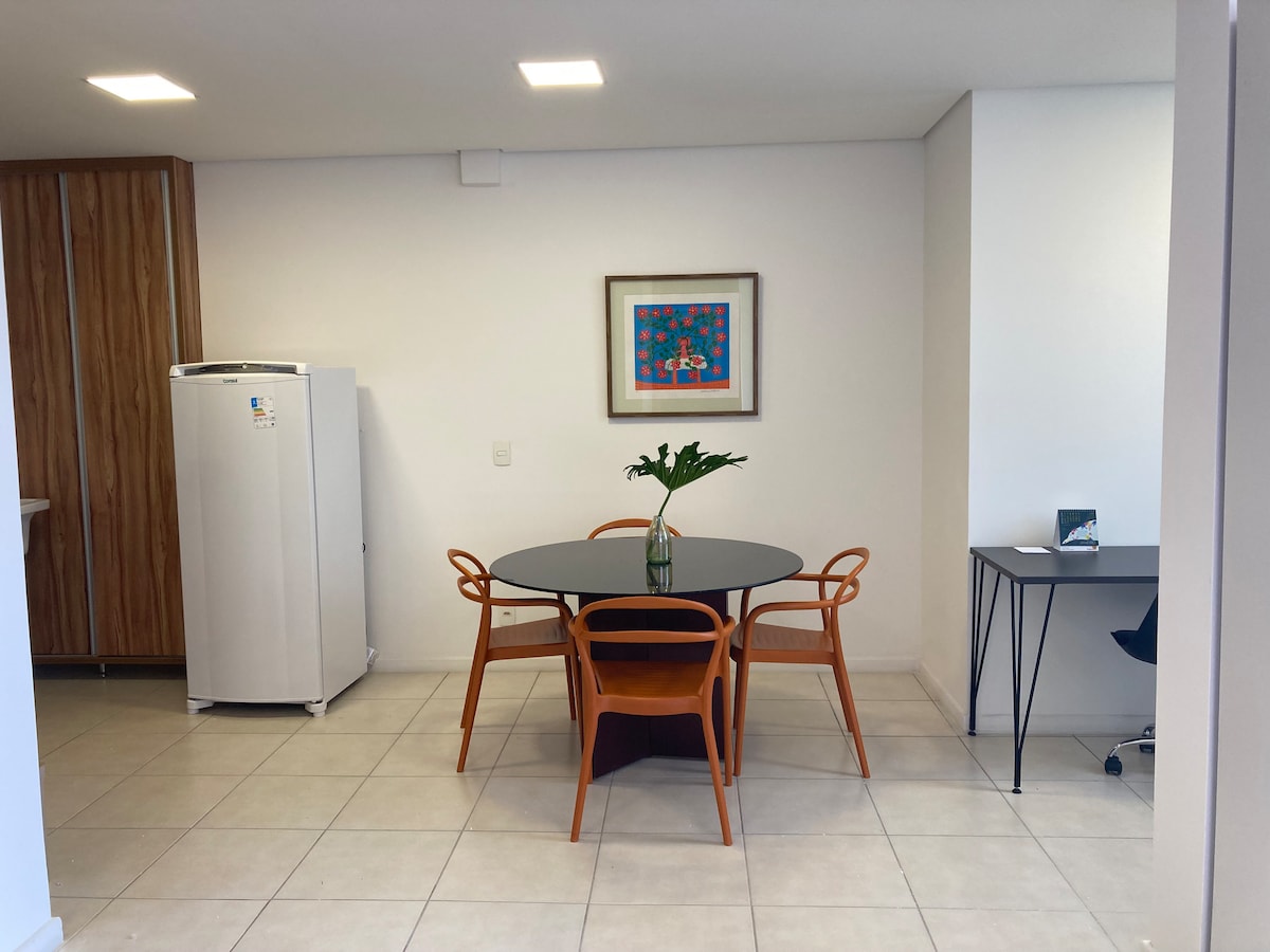 A dining area features a round black table surrounded by four orange chairs. A colorful artwork is displayed on the wall. A white refrigerator stands nearby, while a small workspace with a black desk and chair is visible in the corner.