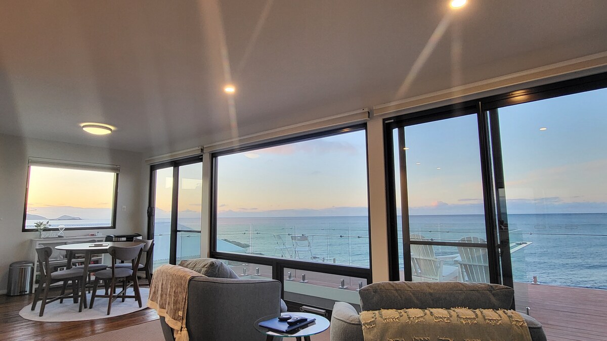 An open living space features large glass windows offering panoramic views of the ocean and sunset. A dining table with wooden chairs is visible, alongside a cozy seating area with light-colored sofas. The warm glow of ceiling lights enhances the welcoming atmosphere.