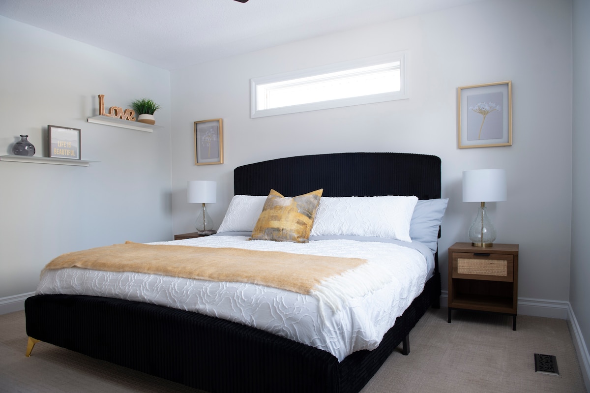 A well-appointed bedroom is presented, featuring a spacious bed adorned with white linens and a golden throw. Two bedside lamps provide soft lighting, while minimal shelving and framed artwork add a touch of decor. A large window allows natural light to fill the room.