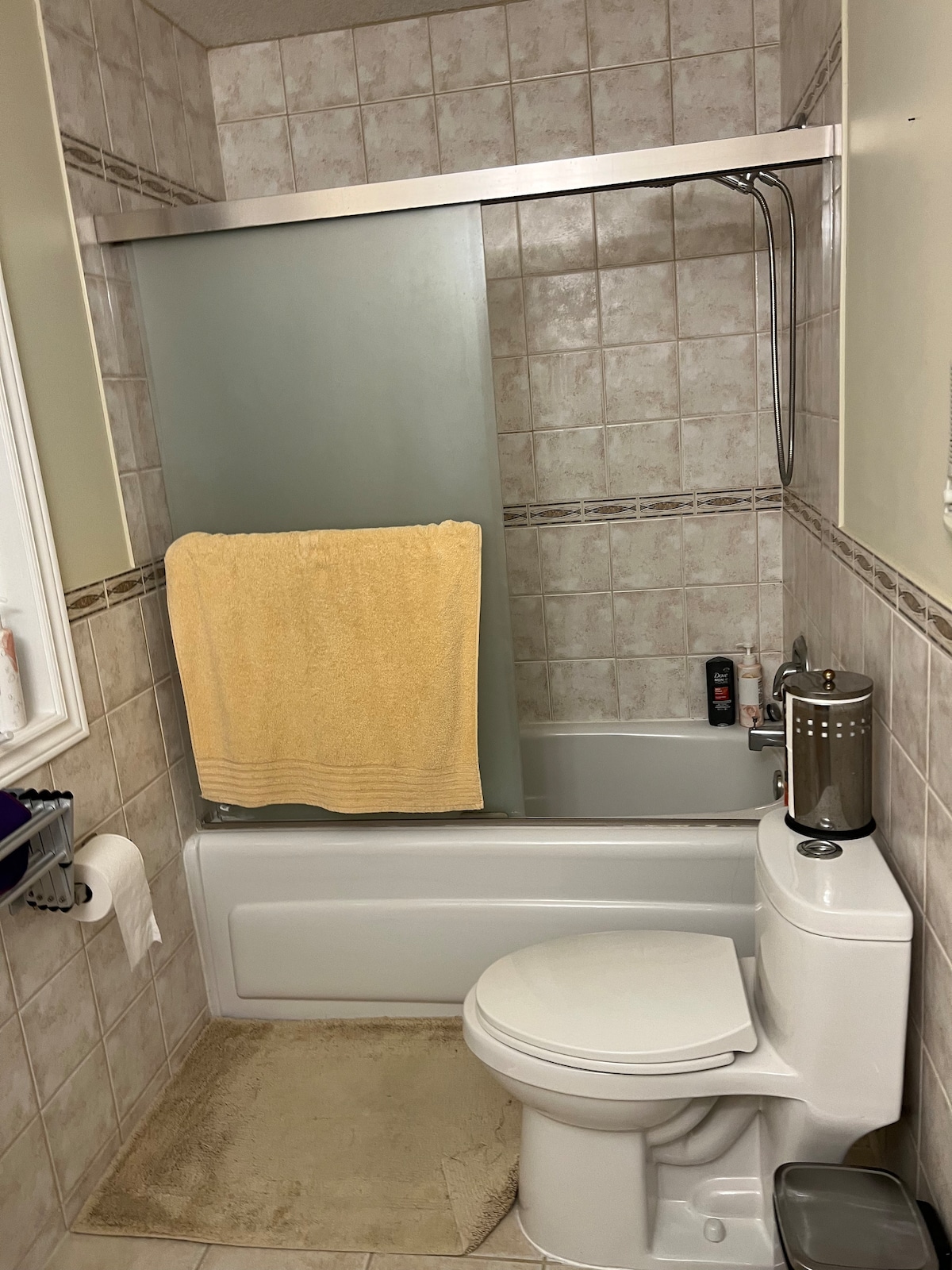 A functional bathroom is visible, featuring a combination bathtub and shower with a clear sliding glass door. A cream-colored towel hangs from a chrome rack, and a small selection of toiletries is placed on the tub ledge. Beige tiles line the floor and walls.