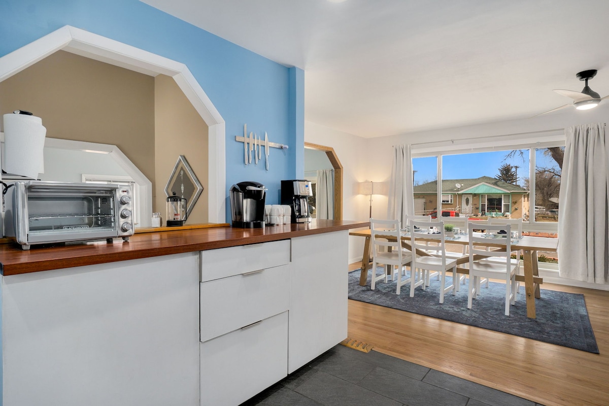 A modern kitchen area is visible, featuring a stylish counter with a microwave and coffee appliances. Light filters through large windows, illuminating a dining space with a white table and chairs. Blue and beige walls create a calm atmosphere.