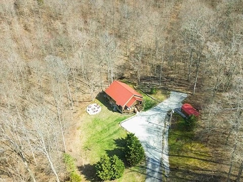 An aerial view showcases a rustic cabin set in a wooded area, surrounded by bare trees. A winding driveway leads to the cabin, which features a red roof. A fire pit is visible in the grassy area beside the cabin.