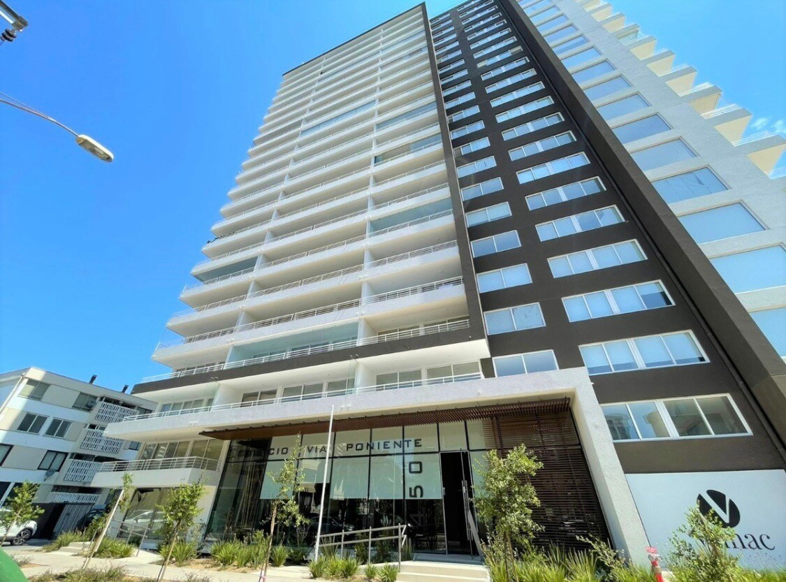 A modern high-rise building is captured from the exterior, showcasing its sleek design and large windows. The entrance features a contemporary facade with the building name displayed prominently. Surrounding greenery enhances the overall appearance, creating an inviting atmosphere in a well-maintained environment.