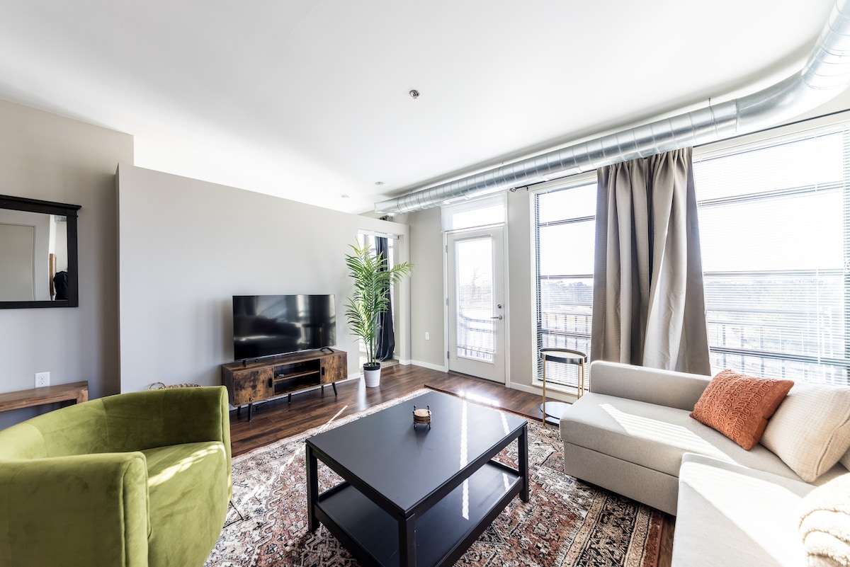 A spacious living room features a 50-inch TV against a light wall, flanked by two large windows with sheer curtains. A green velvet swivel chair and a beige sectional sofa provide comfortable seating around a dark coffee table, while a potted plant adds a touch of greenery.
