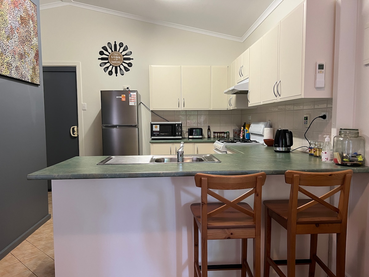 A well-equipped kitchen is visible, featuring light cabinets and a countertop with bar stools. Appliances include a refrigerator, microwave, kettle, and stove. A wall art piece adds visual interest, while the tiled floor enhances the space's functionality.