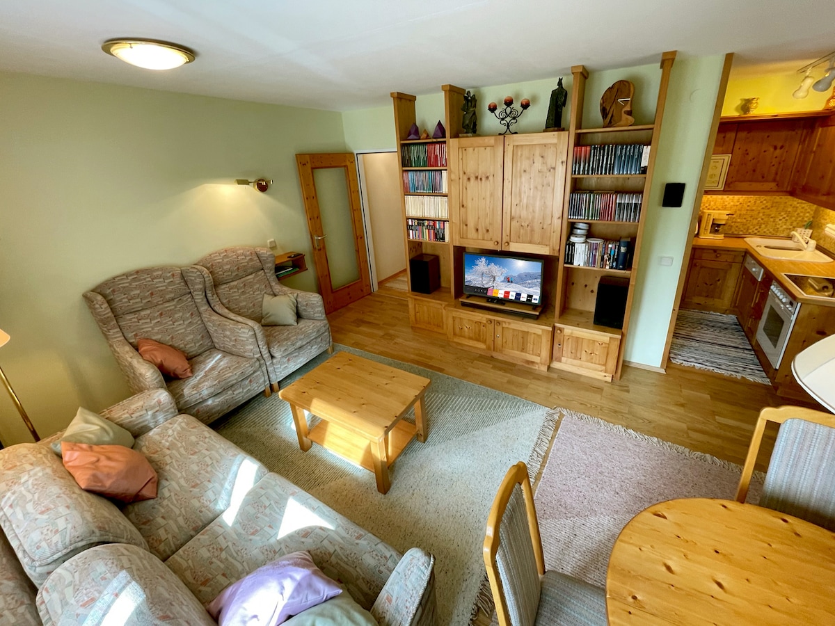 A cozy living area is showcased, featuring a light-toned sofa with throw pillows, a wooden coffee table, and a wooden entertainment unit. Natural light streams in, creating a warm ambiance. The open kitchen area is visible in the background, equipped with essential appliances.