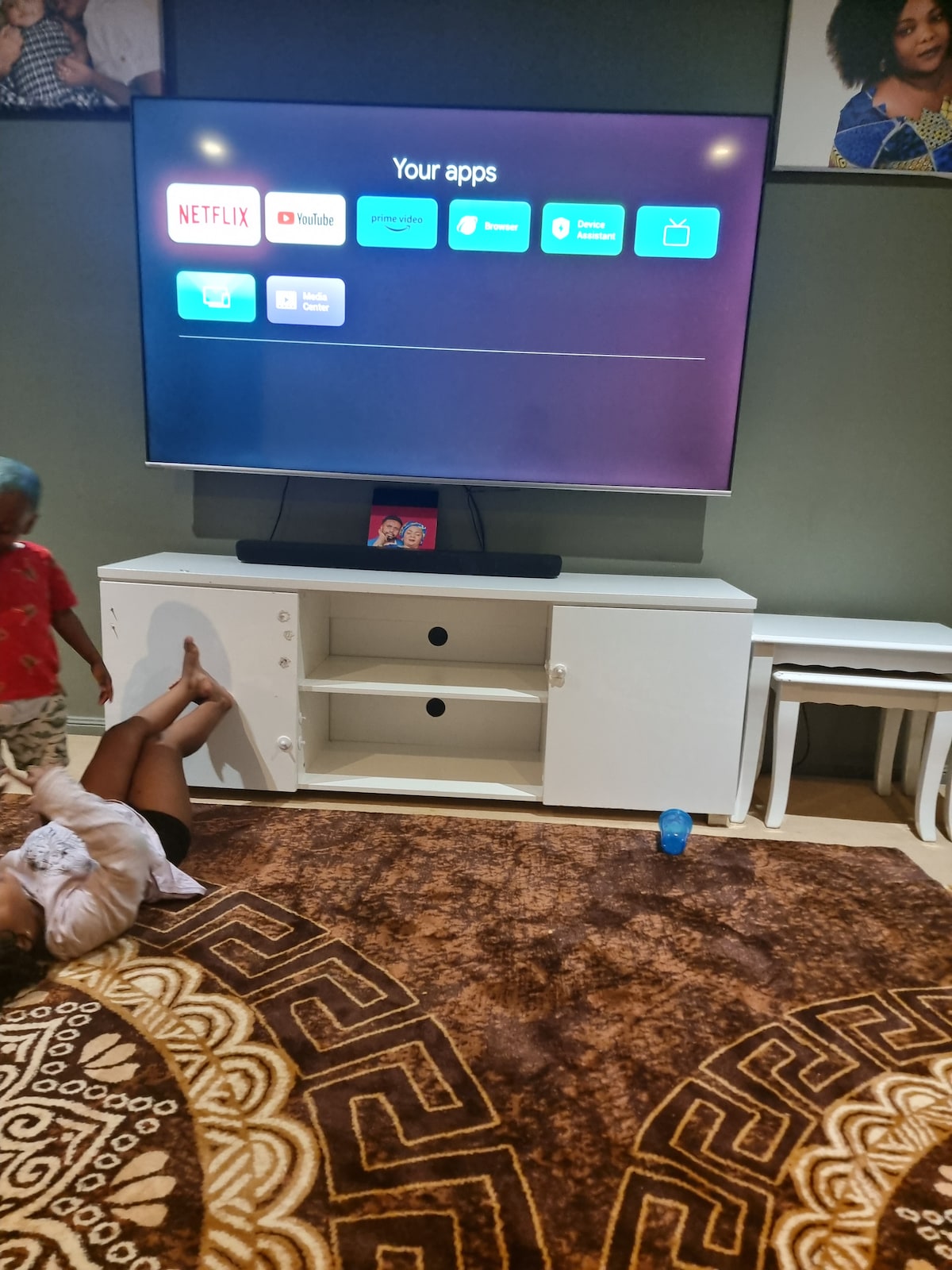 A modern media setup is displayed on a large flat-screen television mounted on a sleek white entertainment unit. The screen shows various app icons, including Netflix and YouTube. A brown patterned rug covers the floor, adding warmth to the space. Two white stools are positioned nearby.
