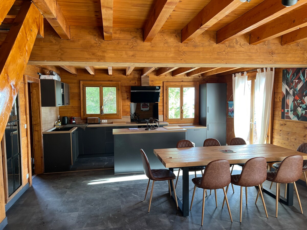 A spacious open-concept living area features wooden beams overhead and a large dining table surrounded by chairs. A modern kitchen is equipped with dark cabinetry and appliances. Natural light filters through multiple windows, creating a bright and inviting atmosphere.