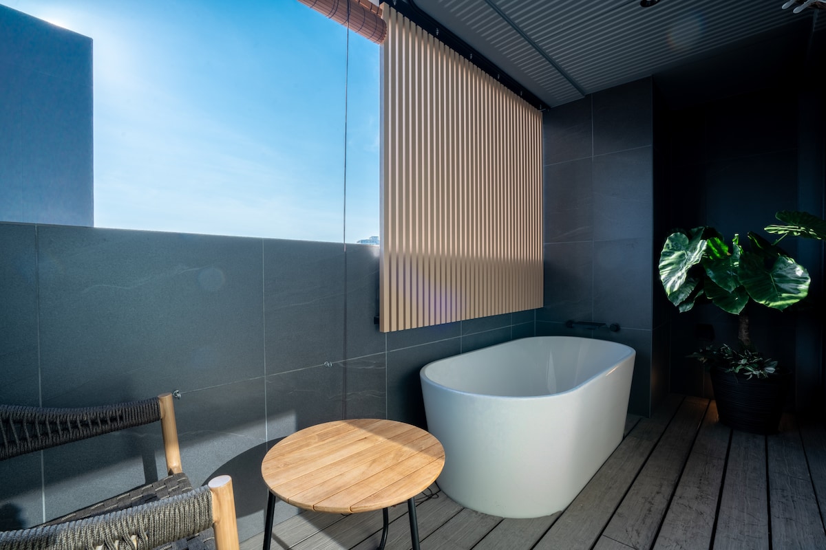A modern bathroom features a freestanding white bathtub positioned beside a window with wooden slatted blinds. A small round wooden table rests nearby, alongside a potted plant. Natural light fills the space, complemented by a minimalist design.
