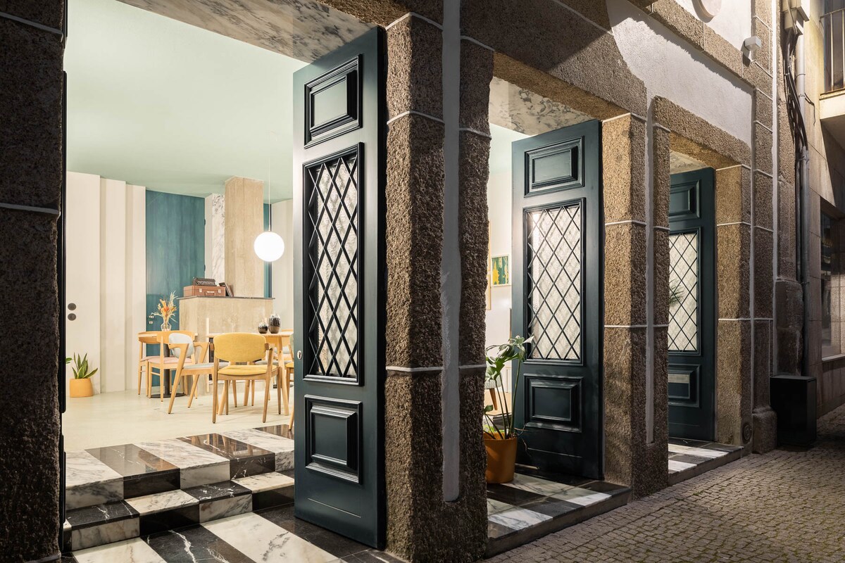 An elegant entrance features three tall, dark wooden doors with diamond-patterned glass panels. The doors open to a well-lit communal area that includes a dining table surrounded by light-colored chairs. The floor is adorned with polished marble tiles, adding a touch of sophistication.