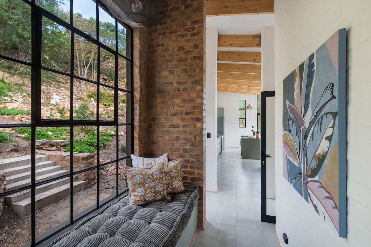 A bright interior space features a large window with black-framed panes, allowing natural light to fill the room. A cozy seating area with patterned cushions is positioned by the window. The visible walls display a blend of natural brick and smooth white finishes.