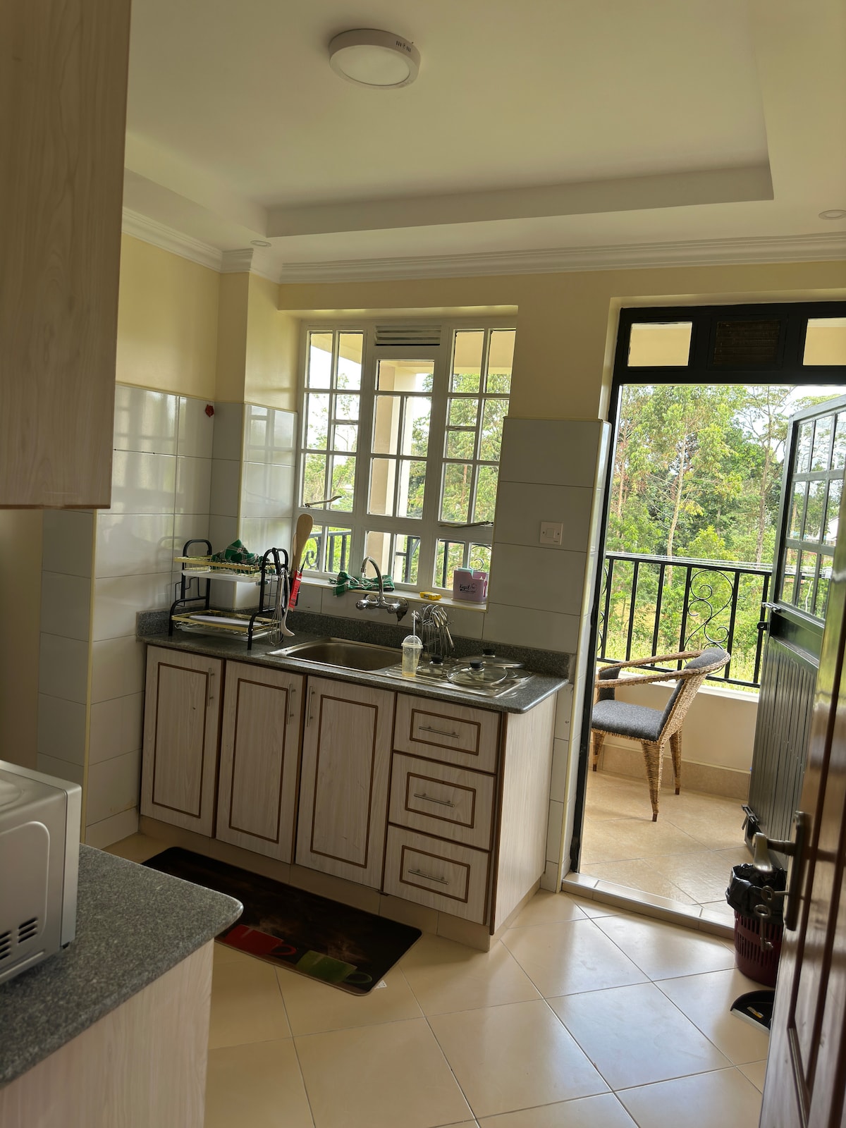A functional kitchen area is highlighted, featuring wooden cabinets and a sink with a dish rack. Natural light fills the space through large windows, with a door leading to a balcony that offers a view of greenery outside.