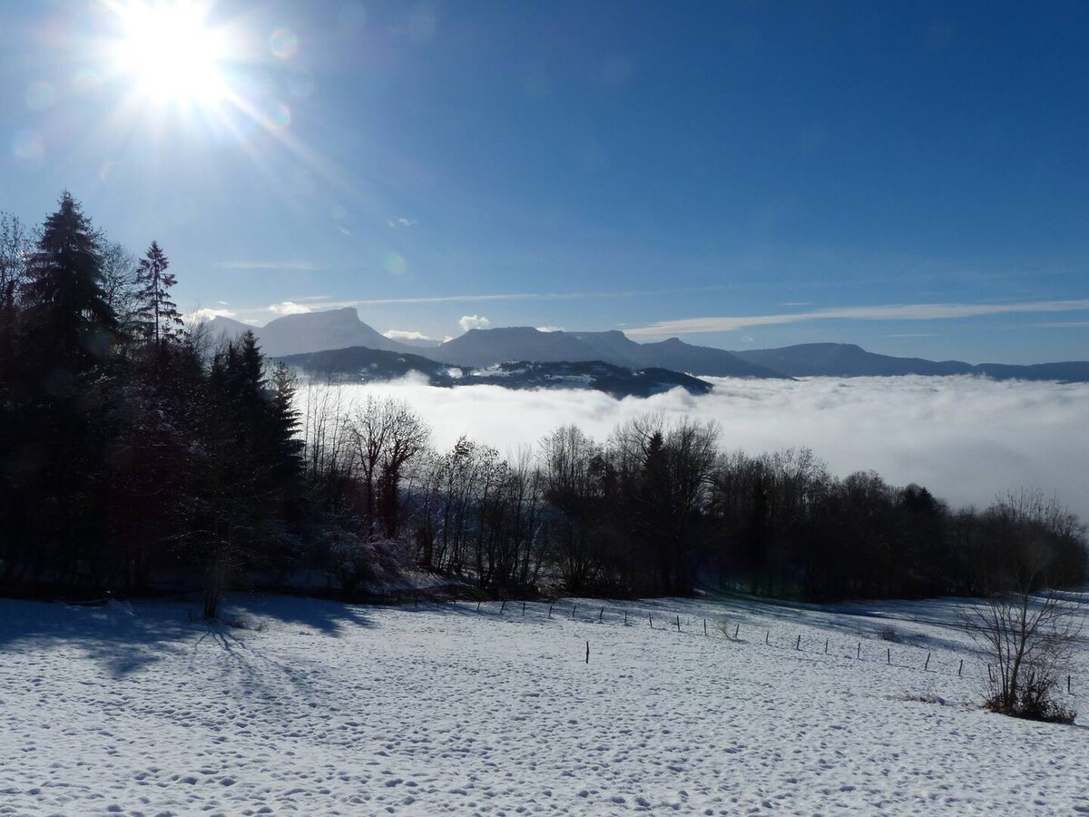 A panoramic view of snow-covered fields is captured under a clear blue sky. Sunlight emanates from the upper left corner, illuminating the distant, mist-blanketed mountains and trees in the foreground, creating a tranquil and serene natural setting.