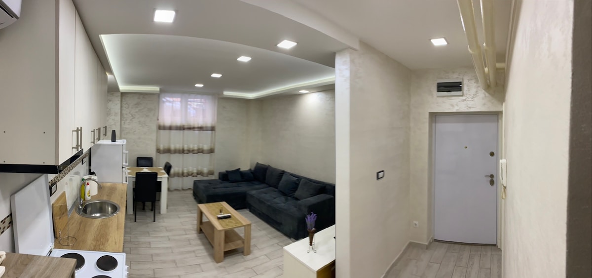 A modern living space is presented with a comfortable dark sectional sofa and a wooden coffee table. Light-colored walls and recessed lighting enhance the ambiance. A small dining area is visible in the background, accompanied by large windows dressed in sheer curtains.
