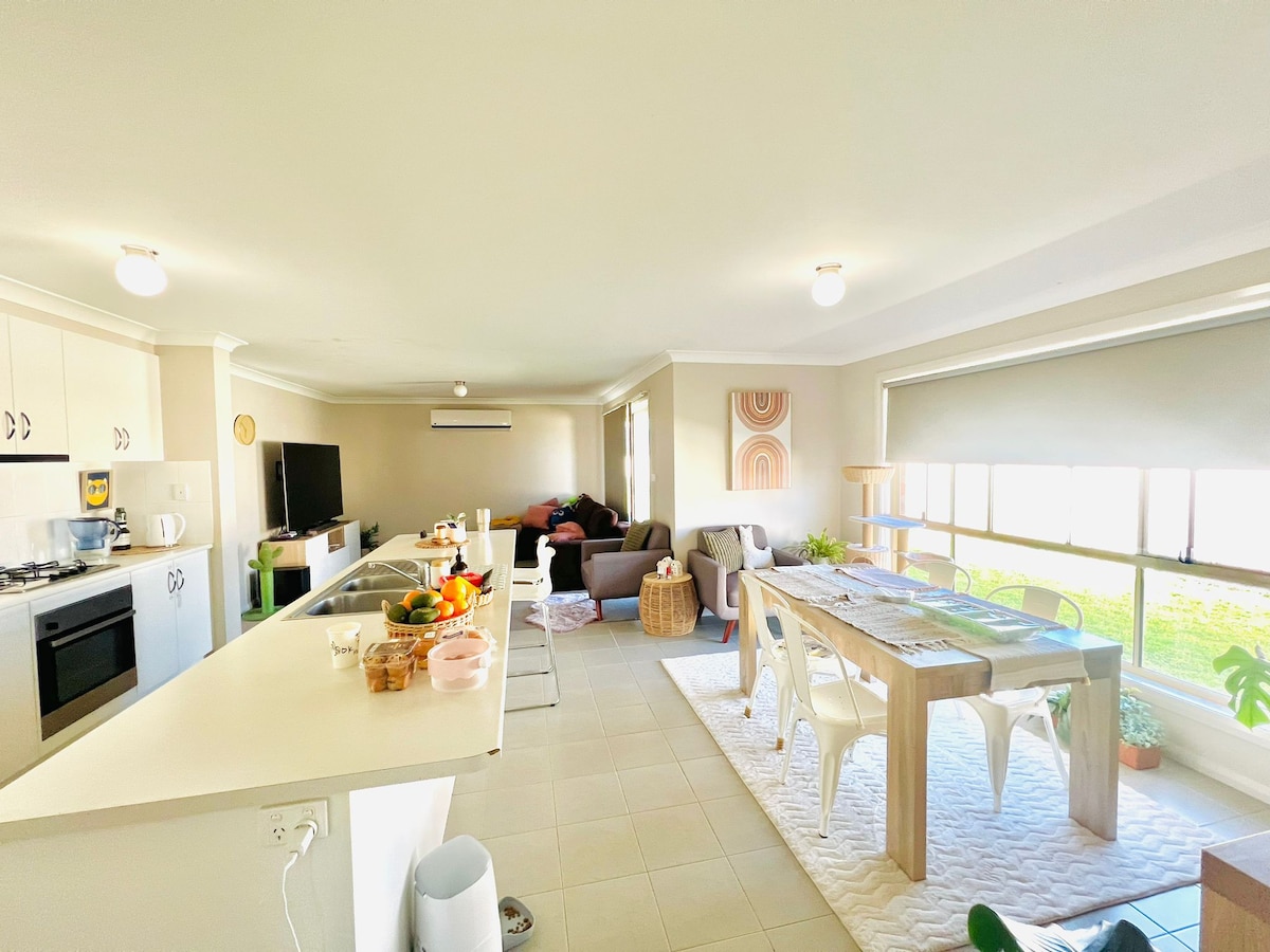 An open living area showcases a kitchen with modern appliances, including a stove and refrigerator, and a dining table with surrounding chairs. A comfortable seating arrangement with soft furnishings is visible, alongside large windows providing natural light and views of the greenery outside.