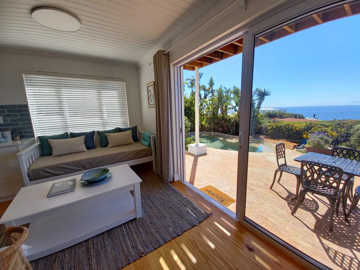 The open-plan lounge features a comfortable seating area with a sofa and large windows allowing natural light. A sliding glass door opens to a spacious patio with a pool, surrounded by greenery and ocean views. A dining table is positioned outside for al fresco dining.