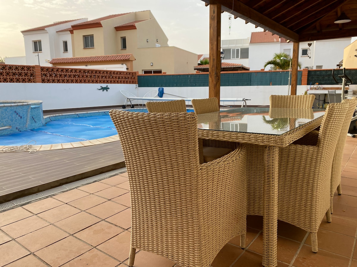 A spacious outdoor dining area is featured, featuring a glass-topped table accompanied by several wicker chairs. The area is shaded by a wooden pergola, with a view of the large pool surrounded by tiled decking and a well-maintained garden.