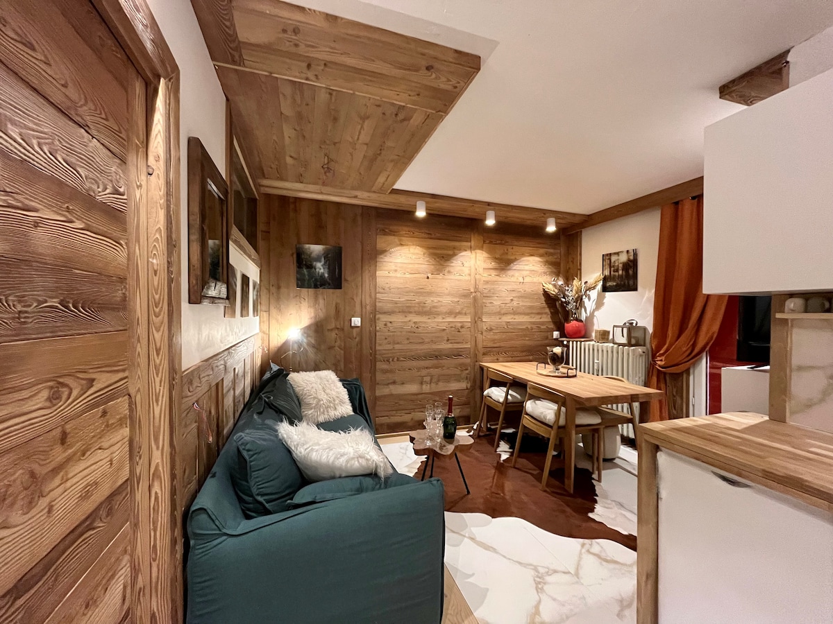 A cozy living area is featured with wooden paneling and warm lighting. A spacious sofa is complemented by a decorative throw. A dining table with chairs occupies the space, along with a few decorative elements on shelves. Natural textures create a harmonious environment.