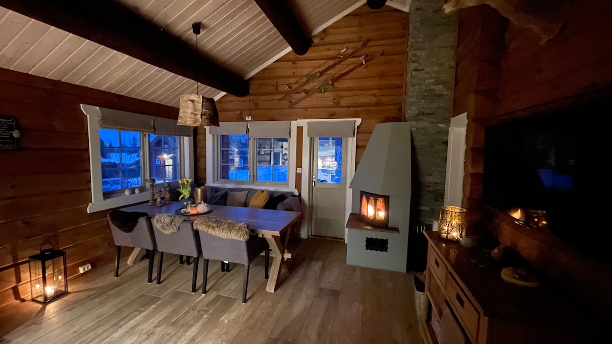 The cozy living area features a large wooden dining table surrounded by four upholstered chairs. A stone fireplace is visible, with a warm flame offering a gentle glow. Windows allow soft twilight light to filter in, complementing the wooden accents and rustic decor.