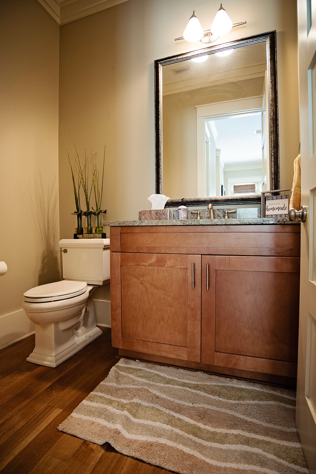 A well-appointed bathroom features a wooden vanity with a granite countertop, paired with a rectangular mirror above. A toilet is positioned near the wall, and decorative plants add a touch of greenery. Soft light is provided by two wall sconces, illuminating the neutral-toned walls.