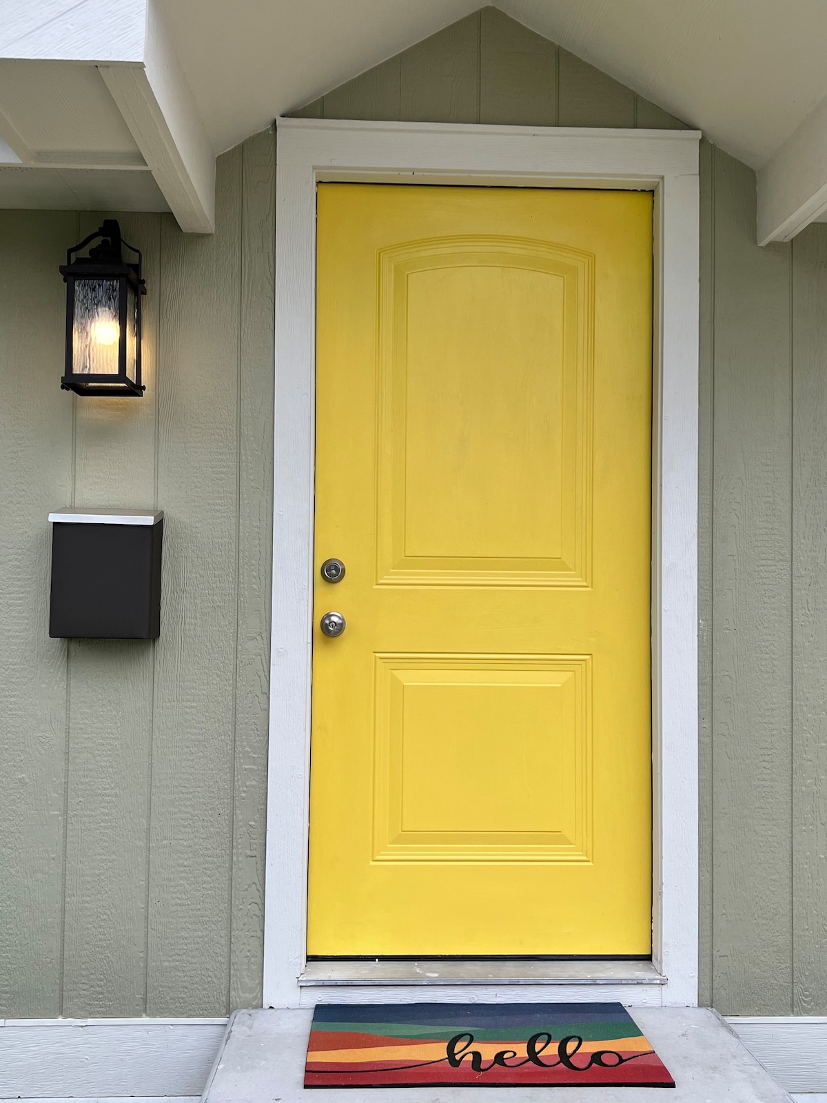 A bright yellow front door is framed by light grey siding. A wall-mounted lantern provides soft lighting beside the door, while a multicolored doormat with the word 'hello' welcomes guests. A mailbox stands nearby, complementing the entrance.