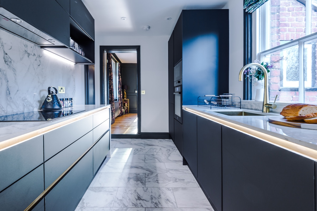 A modern kitchen features sleek black cabinetry and marble countertops, complemented by subtle lighting. The spacious layout includes a stylish sink area with a gold faucet and an oven. A loaf of bread rests on the counter, and light streams in through the nearby window.