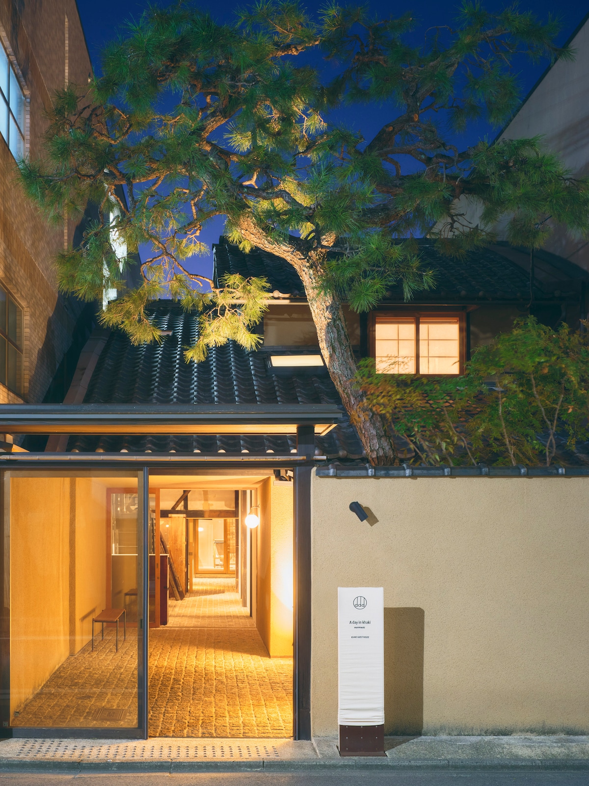 The entrance of the traditional Kyomachiya is highlighted by a pathway illuminated with warm lighting. A mature pine tree stands gracefully beside the building, while the unique roof tiles and soft glow from the windows enhance the welcoming facade.