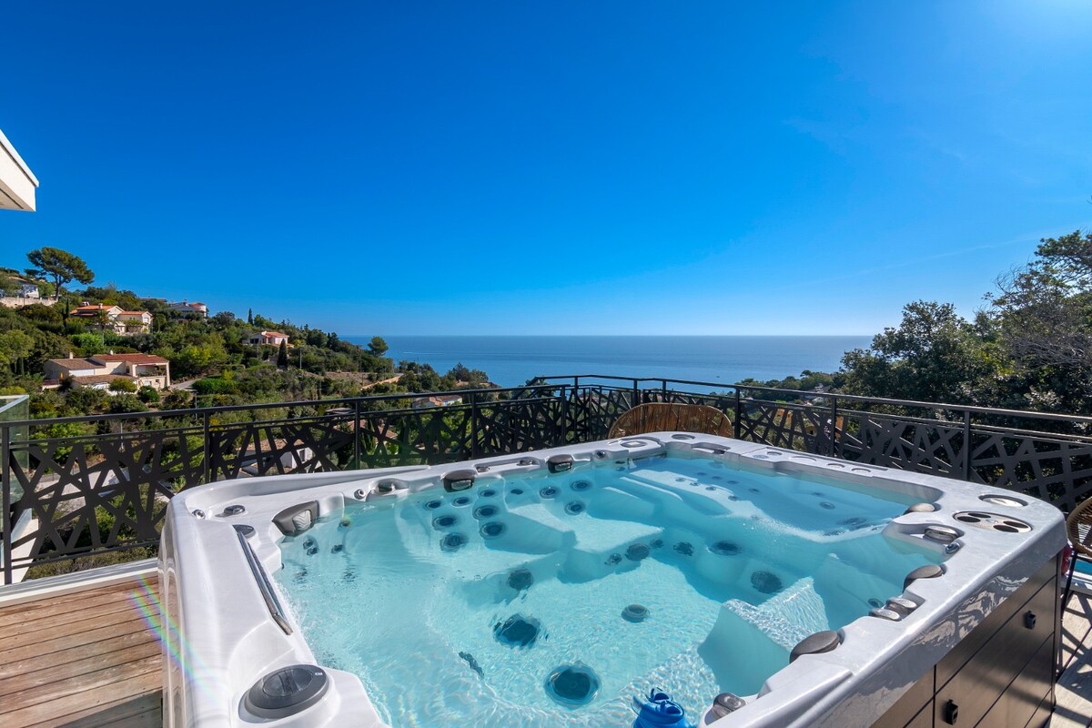 An outdoor jacuzzi is set on a wooden terrace overlooking a panoramic sea view. The hot tub includes seating for eight and features a series of jets. Bright blue skies and lush greenery in the background enhance the serene atmosphere.